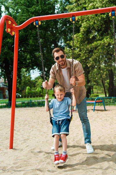 father and son having fun on swing at playground in park and looking at camera