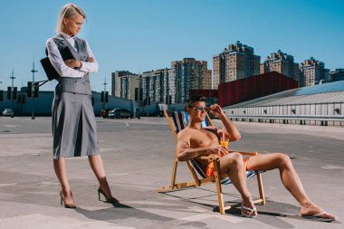 handsome young man relaxing on sun lounger while his lady boss in suit standing next to him with crossed arms on parking clipart