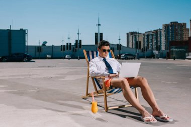 attractive young businessman in shorts working with laptop while sitting on sun lounger on parking clipart