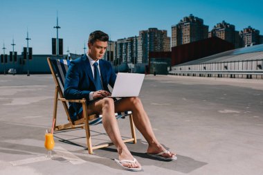 handsome young businessman in shorts and jacket working with laptop while sitting on sun lounger on parking clipart