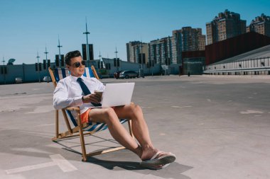 handsome young businessman in shorts working with laptop while sitting on sun lounger on parking clipart