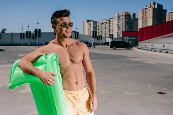 smiling young shirtless man with inflatable bed on parking