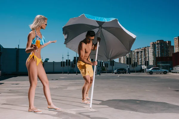 young couple with umbrella in swimsuits tanning on parking