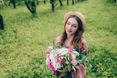 portrait of young smiling woman in hat with bouquet of flowers looking away in park clipart