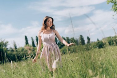 young pensive woman in stylish dress with long hair walking in meadow alone