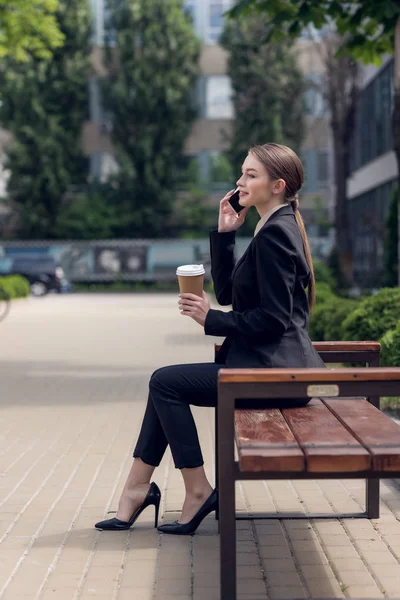 年轻的女商人的侧面视图咖啡去谈智能手机 而在板凳上休息 — 图库照片