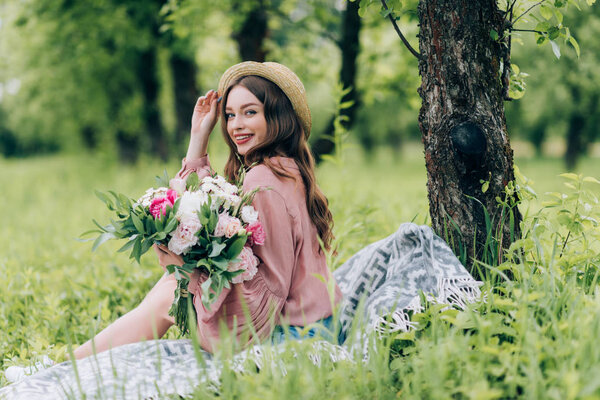 Side view of beautiful smiling woman with bouquet of flowers resting on blanket in park