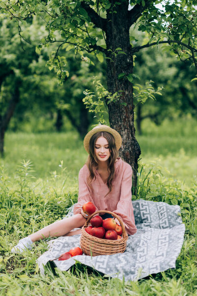 pretty young woman in hat resting on blanket with wicker basket with apples in park