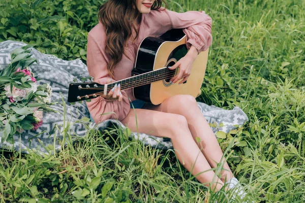 公園の花の花束と毛布で休んでいる間アコースティック ギターを演奏する女性の部分的なビュー  — 無料ストックフォト