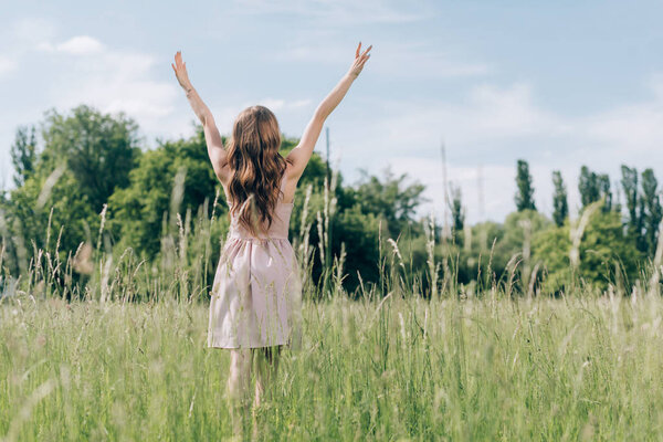 back view of woman in stylish dress with outstretched arms standing in meadow alone