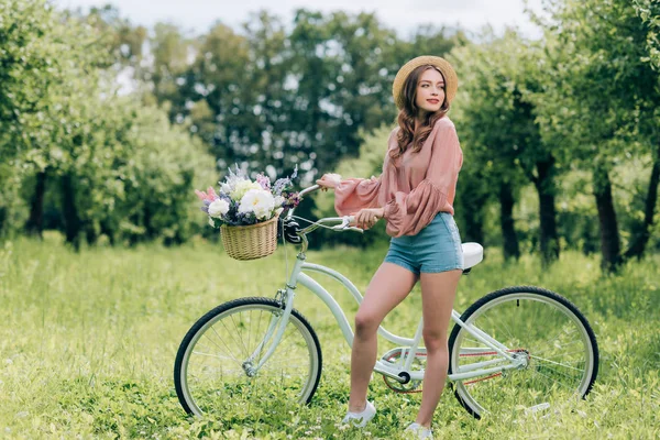 Young beautiful woman standing near retro bicycle with wicker basket full of flowers in forest — Stock Image Young Beautiful Woman Standing Retro Bicycle Wicker Basket Full Flowers Stock Picture