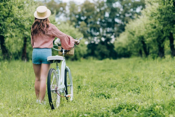 Back view of young woman with retro bicycle with wicker basket full of flowers in forest — Stock Image Back View Young Woman Retro Bicycle Wicker Basket Full Flowers Royalty Free Stock Photos
