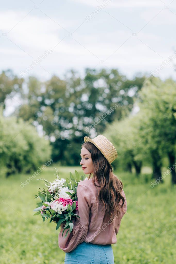 back view of young woman in hat with bouquet of flowers in hands standing in park