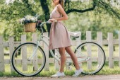 Schnappschuss einer stilvollen Frau im Kleid mit Retro-Fahrrad mit Weidenkorb voller Blumen auf dem Land