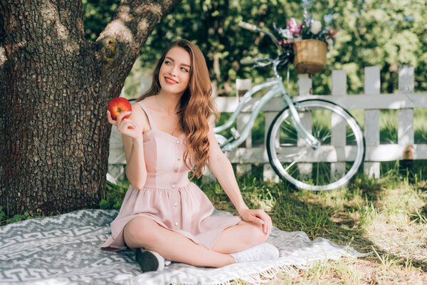 smiling pensive woman with ripe apple in hand resting on blanket under tree at countryside
