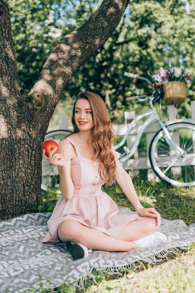 smiling attractive woman with ripe apple in hand resting on blanket under tree at countryside