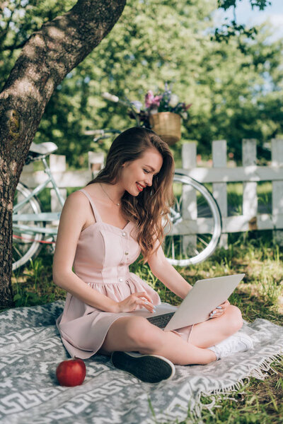 веселая молодая женщина с ноутбуком во время отдыха на одеяле под деревом с велосипедом, припаркованным позади в сельской местности
