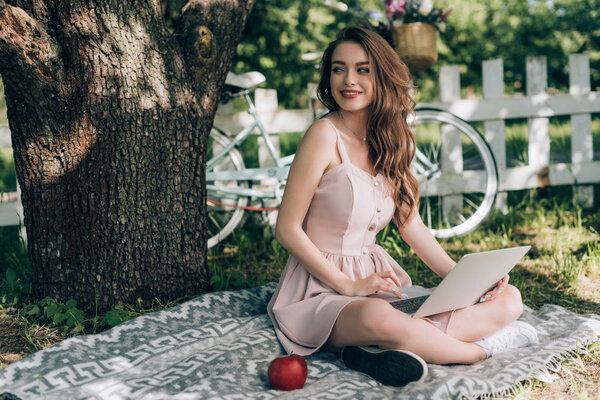 cheerful young woman with laptop resting on blanket under tree with bicycle parked behind at countryside