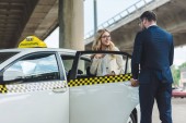 Stylový krasavec otevírání dveří auta, aby usměvavá blondýnka sedí v taxíku