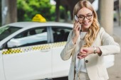 usměvavá blondýnka v brýlích mluví o smartphone a kontrola Náramkové hodinky stoje u taxíku  