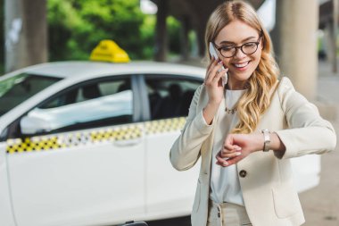 smiling blonde woman in eyeglasses talking by smartphone and checking wristwatch while standing near taxi cab   clipart