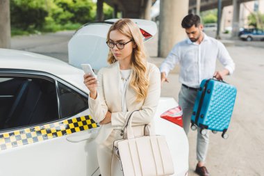 Taksi arabanın bagajında çanta koyarak adam smartphone kullanarak gözlük güzel sarışın kadın  