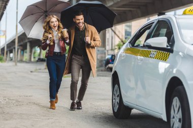 excited young couple with umbrellas running together to taxi cab clipart