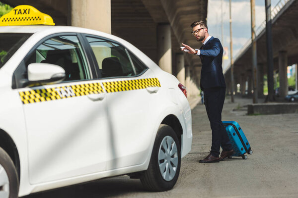 stylish young man with smartphone and suitcase looking at taxi cab