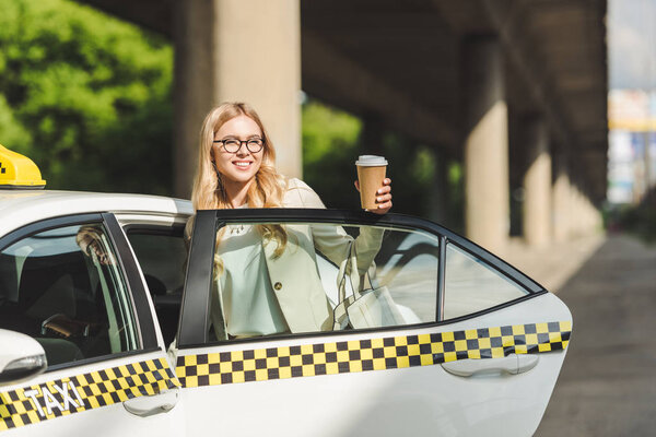 smiling blonde woman in eyeglasses holding coffee to go and looking away while opening door of taxi cab 