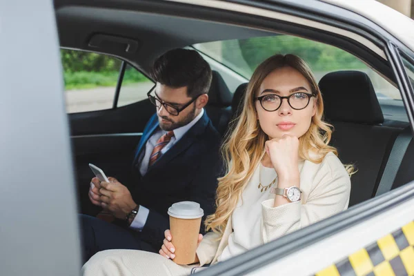 年轻女子拿着纸杯 看着远处的男子使用智能手机在出租车上 — 图库照片