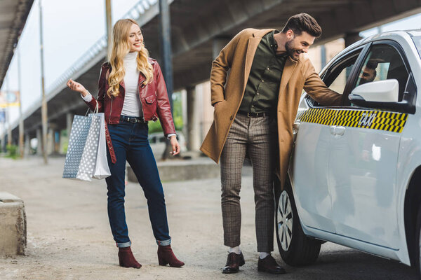 smiling young woman holding shopping bags and looking at happy boyfriend looking into taxi cab