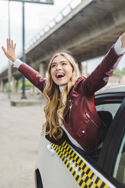 cheerful blonde girl raising hands and looking away through taxi window