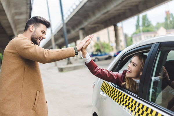 happy young couple giving high five each other, girl sitting in cab and man standing on street 