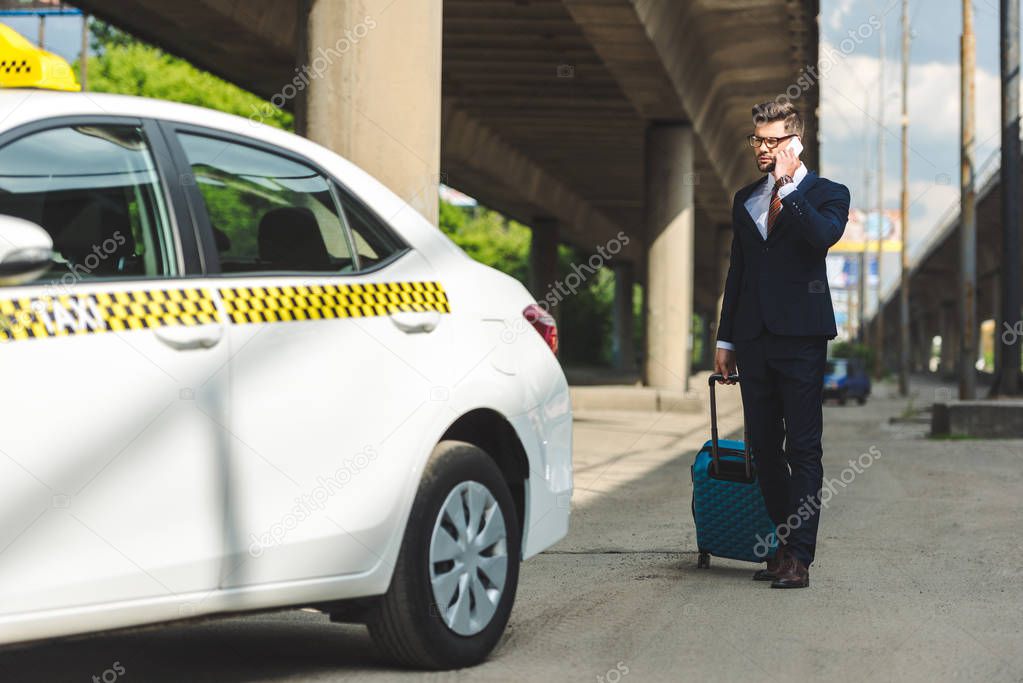 handsome young man in suit and eyeglasses talking by smartphone while standing with suitcase near taxi
