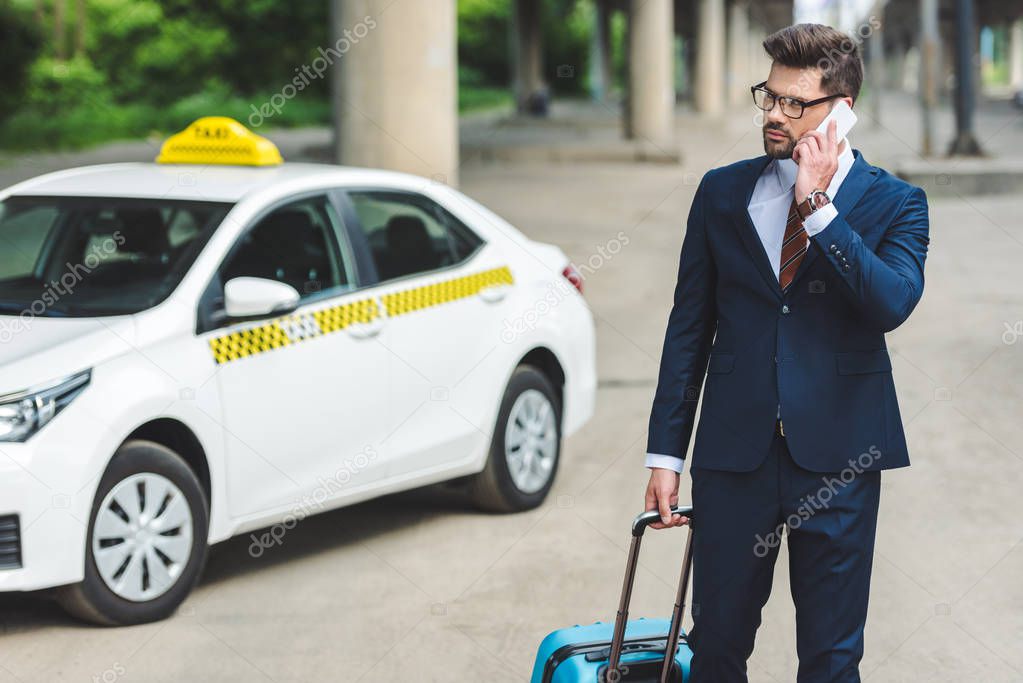 handsome man talking by smartphone while standing with suitcase in taxi cab