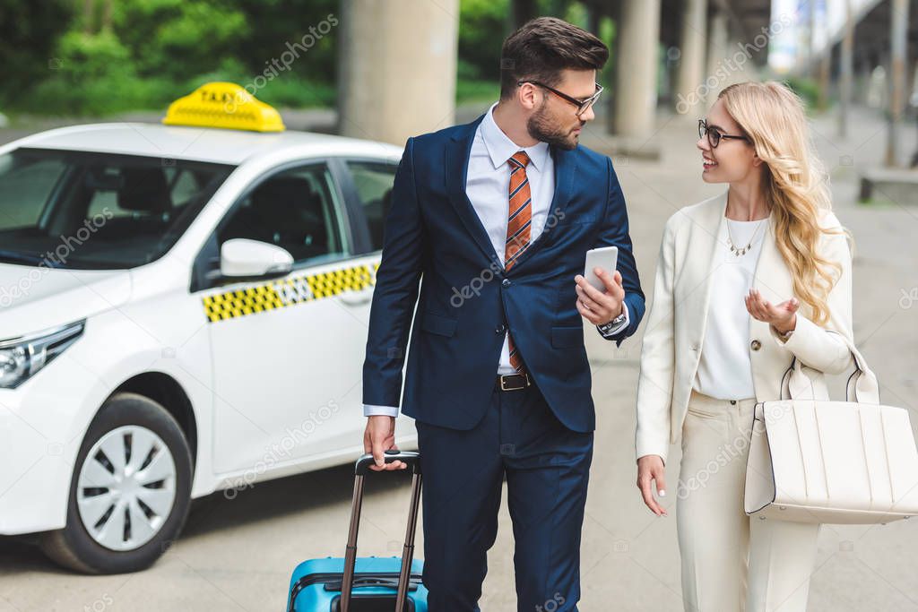 beautiful happy young couple in formal wear smiling each other while going with suitcase near taxi  