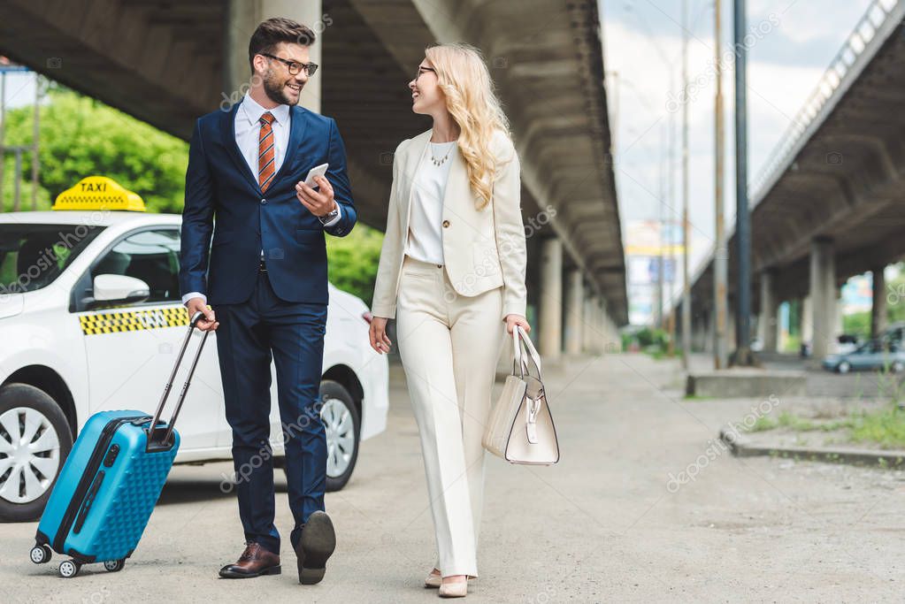 stylish young couple smiling each other while going with suitcase near taxi 