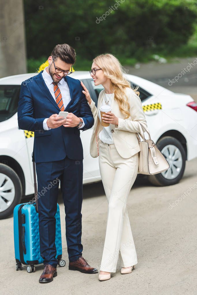 smiling blonde woman holding coffee to go and looking at handsome man using smartphone while standing with suitcase near taxi