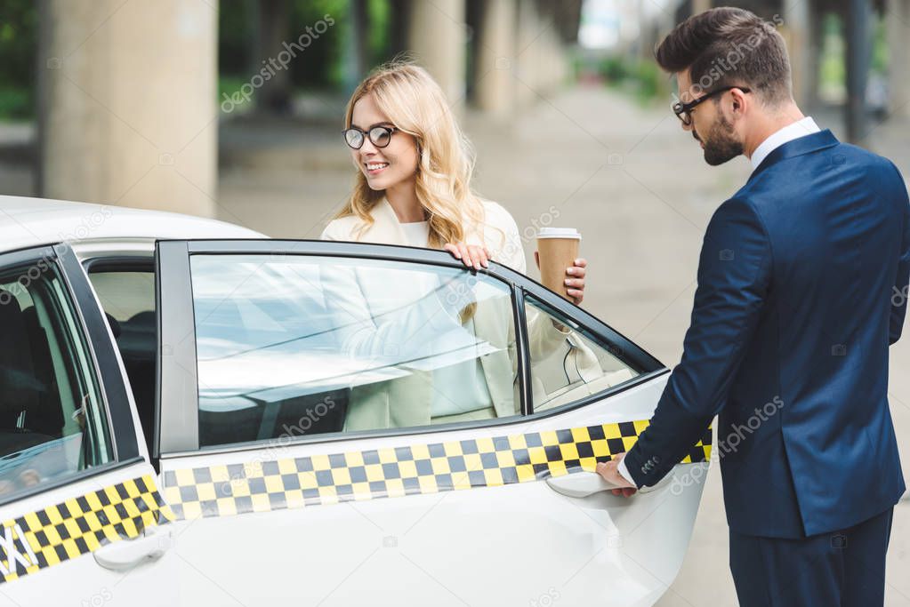 handsome man opening car door while smiling blonde woman with paper cup sitting in taxi
