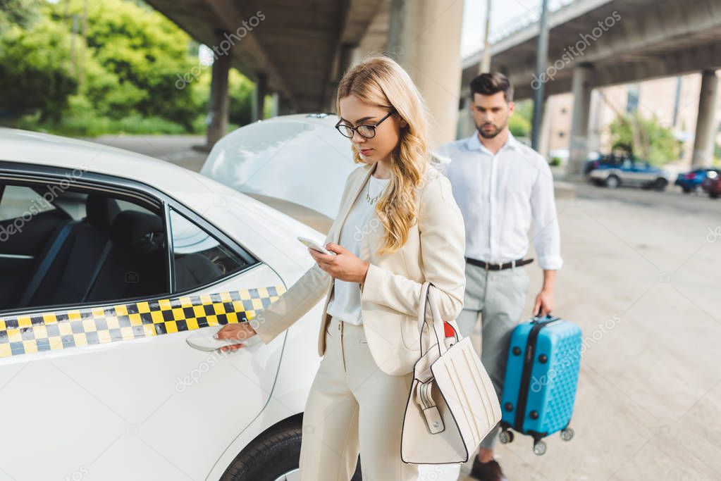 stylish young woman woman using smartphone while man putting suitcase in trunk of taxi 
