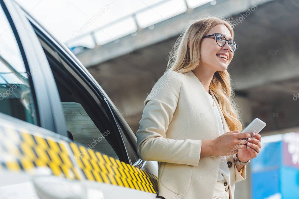 low angle view of smiling blonde girl in eyeglasses holding smartphone and looking away while standing near taxi