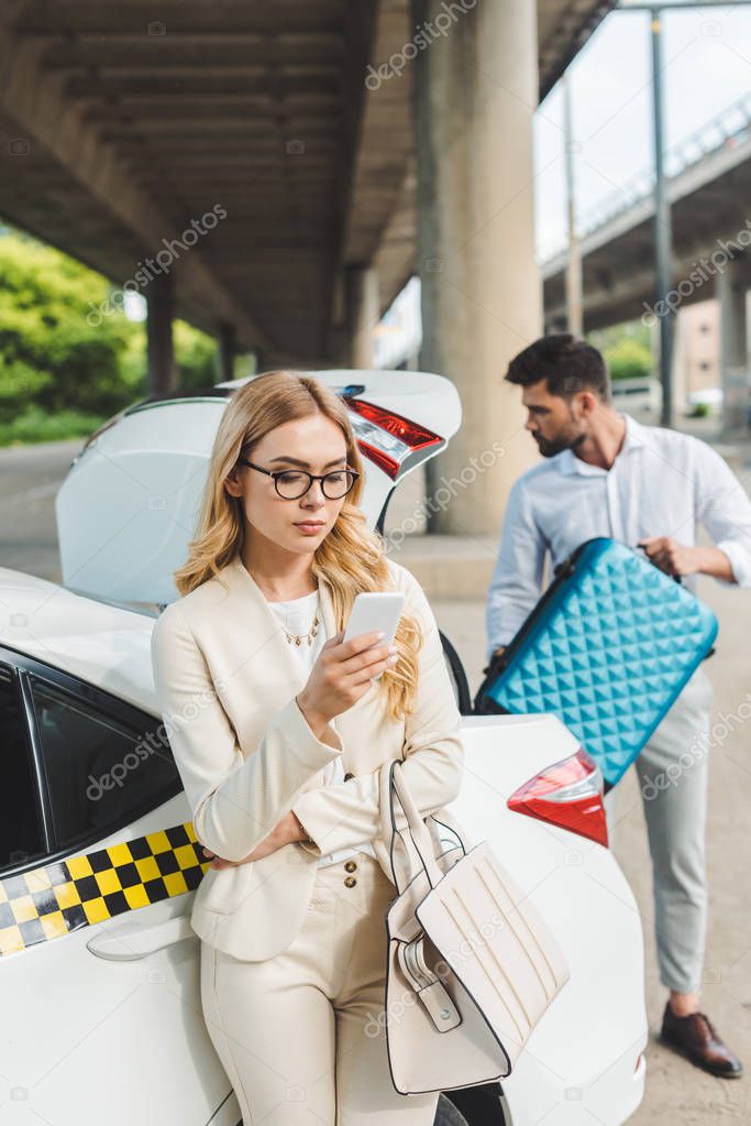 young blonde woman in eyeglasses using smartphone while man putting suitcase in trunk of taxi car 