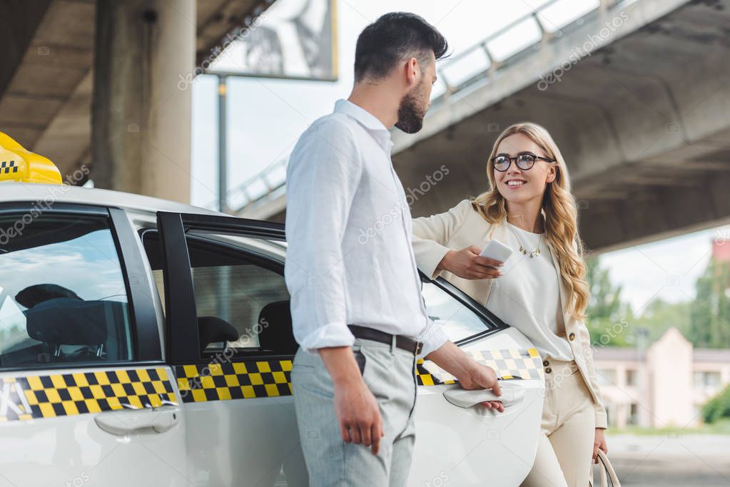 young man opening door of taxi and looking at smiling blonde woman with smartphone