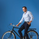 Guapo hombre de negocios sonriente sentado en bicicleta y mirando hacia otro lado en azul