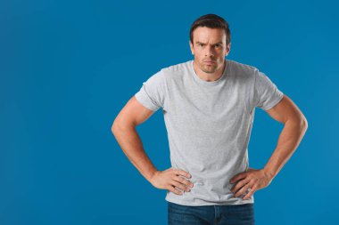frowning man standing with hands on waist and looking at camera isolated on blue clipart