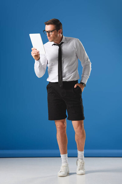 focused man in eyeglasses and shorts using digital tablet while standing with hand in pocket on blue