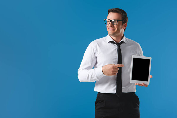 handsome smiling businessman pointing with finger at digital tablet with blank screen and looking away isolated on blue