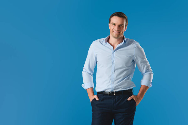 handsome man standing with hands in pockets and smiling at camera isolated on blue