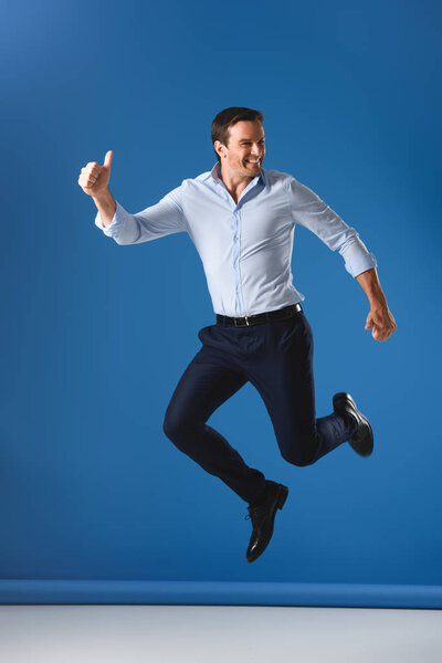 cheerful handsome man showing thumbs up and jumping on blue