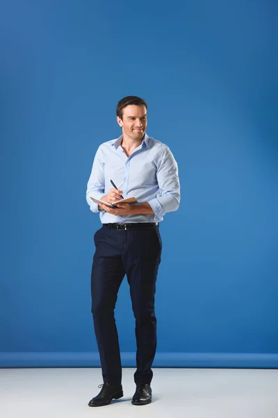 Sonriente Hombre Negocios Escribiendo Cuaderno Mirando Hacia Otro Lado Azul — Foto de stock gratuita